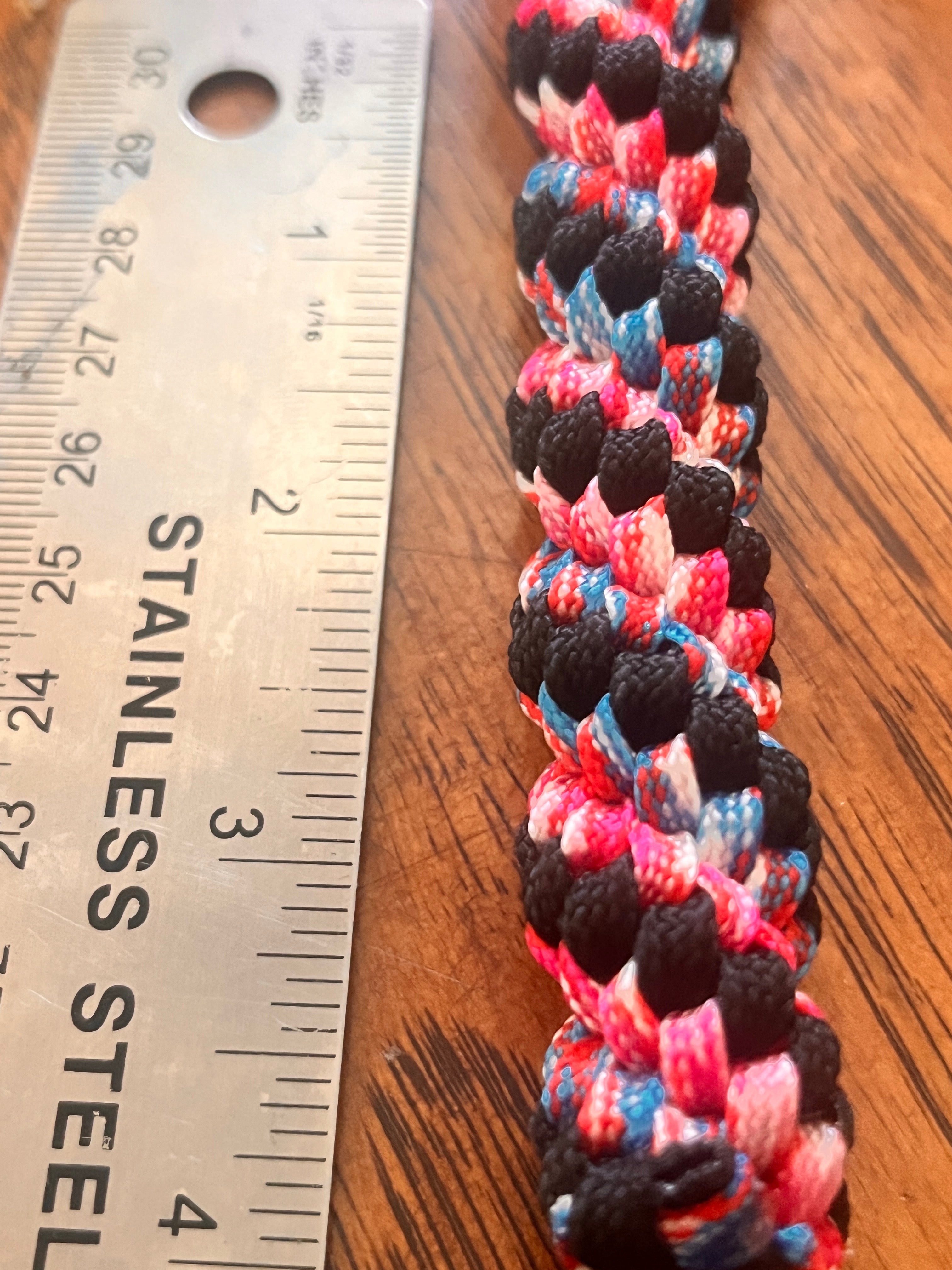 Multicolored braided cord next to a stainless steel ruler on a wooden surface